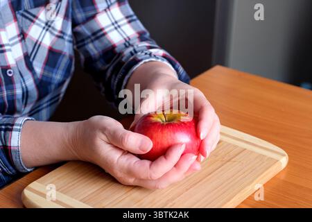 Die Hände einer älteren Frau halten einen roten Reifen, leckeren saftigen Apfel in der Küche an einem braunen Tisch, ohne Gesicht, Nahaufnahme Stockfoto