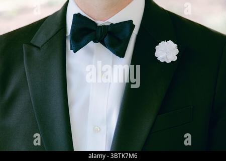 Ein stilvoller Mann im Smoking mit Fliege und weißer Blume steht bei einer Feier im Freien. Stockfoto