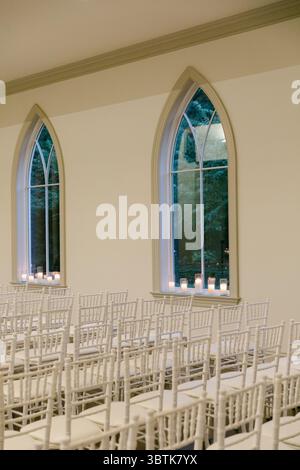 Vor Bogenfenstern sind Reihen weißer Stühle angeordnet, die bei einer Hochzeitszeremonie von Kerzen sanft beleuchtet werden. Stockfoto