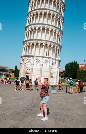 Pisa, Italien - 26. Juni 2025: Eine Frau posiert mit beiden Armen, als würde sie den Schiefen Turm von Pisa hochhalten, eingerahmt von blauem Himmel und Surro Stockfoto