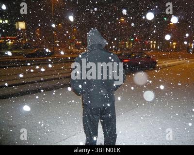 Rückansicht eines Mannes in einer Pufferjacke, der in einer verschneiten Nacht am Straßenrand steht Stockfoto
