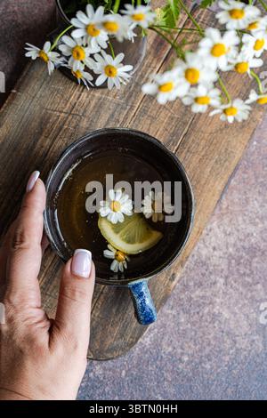 Blick von oben auf die Hand einer Frau, die nach einer Tasse Kamillentee mit Zitrone und Gänseblümchen auf einem hölzernen Schneidebrett und einem Haufen Gänseblümchen greift Stockfoto