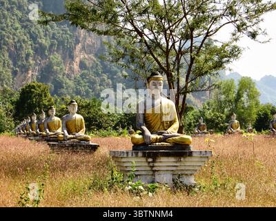 Sitzende Buddha-Statuen im Lumbini Garden, hPa-an, Myanmar, erstrecken sich über trockenes Grasland unter Bergen in einer ruhigen spirituellen Landschaft. Stockfoto