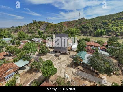 Aus der Vogelperspektive auf ein traditionelles Gemeinschaftshaus (Nhà Rông), das hoch in einem Dorf steht, das vor sanften Hügeln unter hellem Himmel liegt, Kon Tum, Kon Tum, Vietnam. Stockfoto
