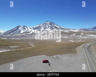 Aus der Vogelperspektive eines einsamen roten Fahrzeugs, das auf dem trockenen Wüstenboden geparkt ist, im Kontrast zu den schneebedeckten Gipfeln der fernen Berge, San Pedro de Atacama, Antofagasta, Chile. Stockfoto
