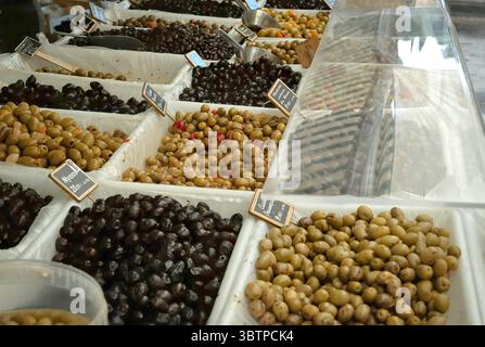 Verschiedene Arten von grünen und schwarzen Oliven werden in offenen Behältern an einem lokalen Marktstand ausgestellt, mit kleinen Schildern gekennzeichnet. Stockfoto
