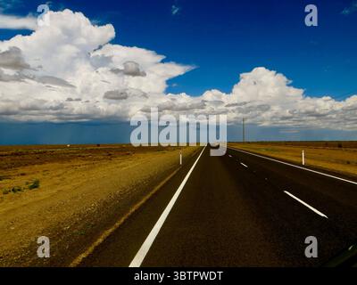 Gerade Autobahn durch das trockene Outback von Western Australia unter einem leuchtend blauen Himmel mit dramatischen Cumulus-Wolken Stockfoto