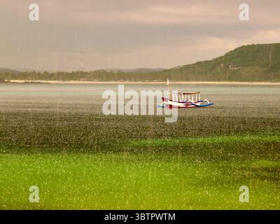 Traditionelles Boot schwimmt auf der regnerischen Meeresoberfläche in der Nähe von Lombok, Indonesien, mit nebeliger Küste im Hintergrund Stockfoto