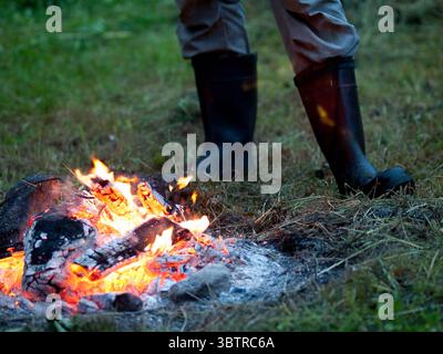 Helles Lagerfeuer, das mit glühenden Baumstämmen und Gluten in einem Wald brennt, während eine Person in der Nähe steht – ein warmer Moment in der Wildnis. Stockfoto