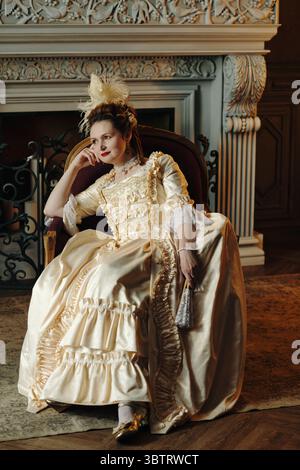 Kaukasische junge Erwachsene Frau, die in einem kunstvollen Barockkleid sitzt, Kopf auf der Hand liegend und im Renaissance-inspirierten Innenraum nachdenklich zur Seite blickt Stockfoto
