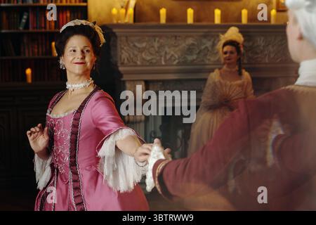 Kaukasische Frau mittleren Alters im barocken Stil, die mit einem kaukasischen Mann mittleren Alters im historischen Innenraum tanzt, eine weitere kaukasische Frau mittleren Alters im Hintergrund Stockfoto
