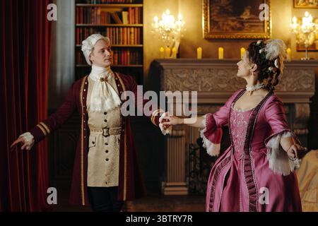 Junger Mann und Frau in barocken Kostümen, die elegante Tänze in kunstvoll verzierten Innenräumen im Renaissance-Stil mit Bücherregalen und Kamin aufführen Stockfoto