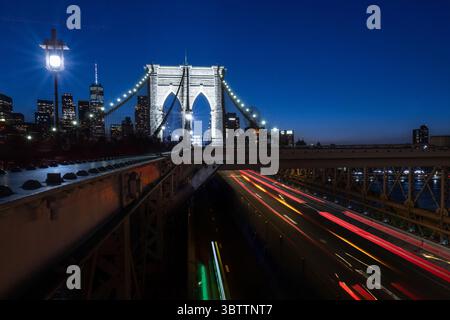 Brooklyn Bridge Traffic - Autos bei Sonnenuntergang Abend und Einbruch der Dunkelheit - Zeitverzögerungsbewegung verschwimmen Steinbögen in Lower Manhattan New York City mit Skyline Stockfoto