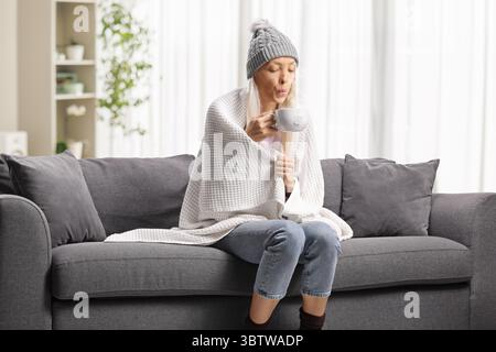 Eine kranke Frau, in eine Decke gewickelt, sitzt auf einem Sofa und hält eine Tasse heißes Getränk Stockfoto