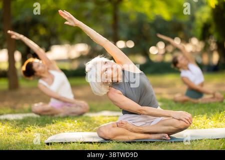 Sportliche alte Dame sitzt auf einer Sportmatte und macht Yoga-Meditation in Lotusposition und Hände hoch mit einer Gruppe von Frauen zusammen im grünen Park tagsüber Stockfoto