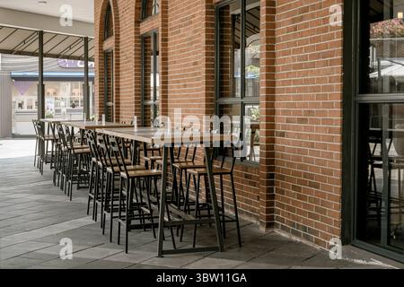 Jakarta, Indonesien, 15. Juli 2025. Sitzbereich auf der Außenterrasse eines Cafés mit hohen Holztischen und schwarzen Metallhockern neben einer roten Ziegelwand. Stockfoto