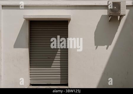 Eine geschlossene graue Rollladentür an einer weißen Wand mit Klimaanlage, die bei hellem Sonnenlicht starke Schatten erzeugt. Minimalistisches urbanes Detail. Stockfoto