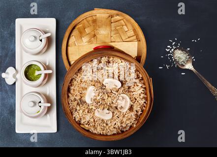 Aromatischer Pilzreis, serviert in einem Bambusdampfer, garniert mit Pilzscheiben. Begleitet von einem Trio tauchender Saucen auf einem Keramiktablett, b Stockfoto