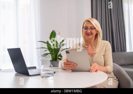 Professionelle Frau mittleren Alters in angenehmer Atmosphäre, die Online-Beratung anbietet, Notebook und Tablet für die Unterstützung des psychischen Wohlbefindens verwendet. Stockfoto