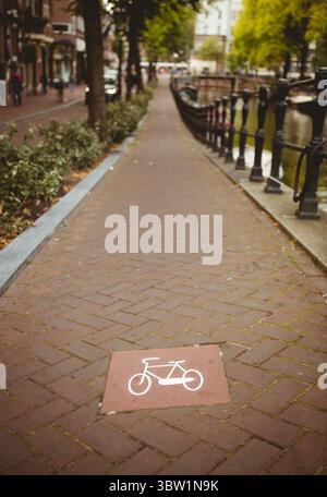 Die rote Kachel mit dem Fahrrad-Symbol markiert einen flachen Fahrradweg entlang eines Backsteinkanals mit Geländer Stockfoto