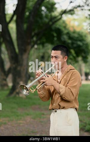 Männlicher Musiker, der sich auf das Spielen einer silbernen Trompete in einem friedlichen Garten konzentrierte Stockfoto