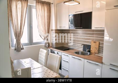 Diese moderne, minimalistische Küche verfügt über elegante weiße Schränke, ein kompaktes Layout und viel natürliches Licht aus dem Fenster, was eine helle und funktionelle Atmosphäre schafft Stockfoto