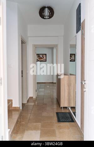 Moderner Flur mit Holzakzenten und Fliesenboden, minimalistischer Einrichtung mit weißen Wänden, Tageslicht und eleganter Ästhetik. Stockfoto