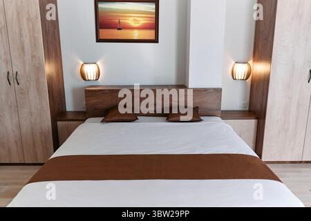 Ein modernes Schlafzimmer mit einem eleganten Bett in neutralen Farben, ergänzt durch Holzmöbel und heitere Wandbilder, die eine gemütliche und einladende Atmosphäre schaffen Stockfoto