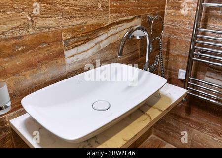 Ein modernes Badezimmer mit einem eleganten weißen Waschbecken mit Chromhahn, montiert auf einer Marmorplatte, auf braunen strukturierten Fliesen und einem polierten t t Stockfoto
