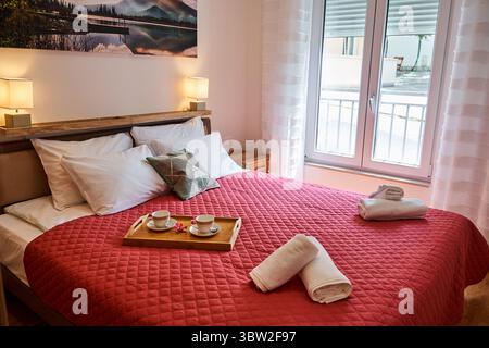 Ein gemütliches Schlafzimmer mit roter Steppdecke, einem ordentlich angeordneten Bett, natürlichem Licht durch ein großes Fenster und einem Holztablett mit Tassen, die eine ruhige und ruhige Atmosphäre schaffen Stockfoto