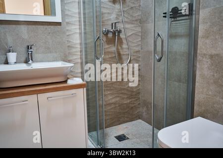 Modernes Bad mit Glasdusche, stilvollem weißem Waschbecken, Kosmetikbereich mit Holzakzenten und eleganten Fliesen für einen eleganten, modernen Look. Stockfoto