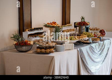 Ein üppiges Buffet mit Ständen, an denen Gebäck, frische Obstspieße und herzhafte Snacks serviert werden, steht in einem dezent beleuchteten Saal für eine Veranstaltung bereit Stockfoto