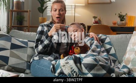 Kaukasische Familie zu Hause Medizin Gesundheit Pandemie Mutter Kuscheln mit Tochter Temperatur überprüfen mit Thermometer hohes Fieber ungesundes krankes Kind Stockfoto