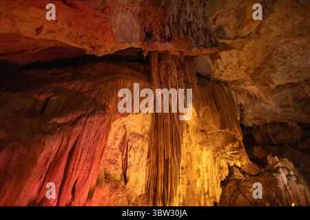 Besucher bestaunen beeindruckende Felsformationen in einer Kalksteinhöhle, die von orangefarbener und gelber Beleuchtung beleuchtet wird und die komplizierten Details der Sta hervorhebt Stockfoto