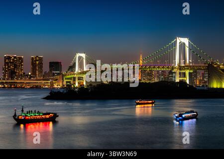 Blick auf beleuchtete Boote, die auf dunklem Wasser unter einer hell beleuchteten Brücke segeln, und auf die Skyline der Stadt unter einem Abendhimmel, Tokio, Japan. Stockfoto