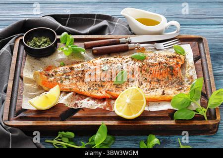 Gegrillte ganze Lachsfilets mit Haut auf einem rustikalen Holzschneidebrett mit frischem Basilikum, Zitronenscheiben, Besteck und Gewürzen auf Holztisch, Horizo Stockfoto