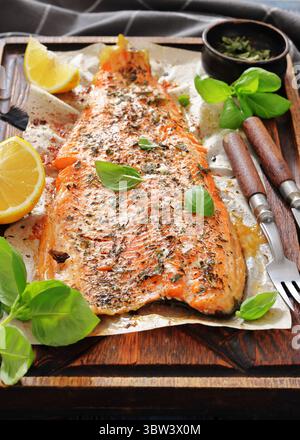 Gegrillte ganze Lachsfilets mit Haut auf einem rustikalen Holzschneidebrett mit frischem Basilikum, Zitronenscheiben, Besteck und Gewürzen auf Holztisch, vertica Stockfoto