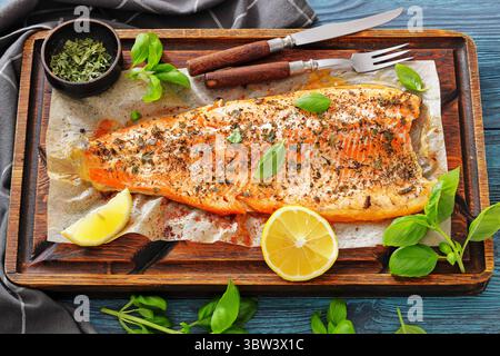 Gegrillte ganze Lachsfilets mit Haut auf einem rustikalen Holzschneidebrett mit frischem Basilikum, Zitronenscheiben, Besteck und Gewürzen auf Holztisch, Horizont Stockfoto