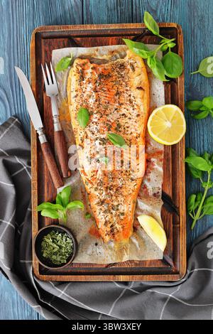 Gegrillte ganze Lachsfilets mit Haut auf einem rustikalen Holzschneidebrett mit frischem Basilikum, Zitronenscheiben, Besteck und Gewürzen auf Holztisch, vertica Stockfoto