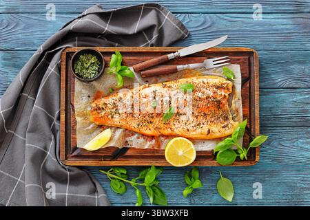 Gegrillte ganze Lachsfilets mit Haut auf einem rustikalen Holzschneidebrett mit frischem Basilikum, Zitronenscheiben, Besteck und Gewürzen auf Holztisch, flaches La Stockfoto