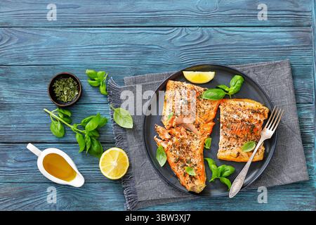 Gegrillte ganze Lachsfilets mit Haut auf einem Teller mit frischem Basilikum, Zitronenscheiben, Gabel auf Holztisch, horizontale Ansicht von oben, flache Lage, close-u Stockfoto