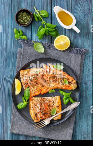 Gegrillte ganze Lachsfilets mit Haut auf einem Teller mit frischem Basilikum, Zitronenscheiben, Gabel auf Holztisch, vertikale Ansicht von oben, flache Lage, Nahaufnahme, Stockfoto