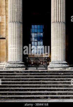 Eine architektonische Aufnahme eines Gebäudeeingangs mit zwei großen Säulen, einer Treppe und einem Fenster, das den Himmel reflektiert. Stockfoto