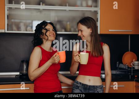 Lächelnde Mutter und Tochter im Teenageralter in roten Tops genießen gemeinsam in der Küche Morgenkaffee und teilen einen unbeschwerten Moment, der Freude und Zuneigung fängt Stockfoto