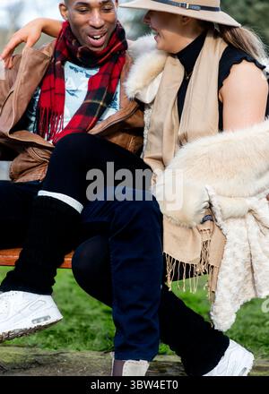 Ein Paar sitzt auf einer Bank, lächelt und lacht. Der Mann trägt eine braune Lederjacke und einen roten Schal, während die Frau einen Hut und trägt Stockfoto
