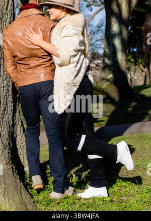 Ein Paar umarmt sich in der Nähe eines Baumes in einem Park. Die Frau trägt einen Hut, einen weißen Pelzmantel, schwarze Leggings und weiße Turnschuhe. Der Mann trägt einen br Stockfoto