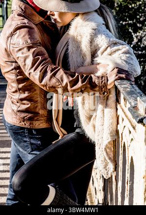 Ein Paar umarmt sich auf einer Brücke, der Mann in einer Lederjacke und die Frau in einem Pelzmantel und Hut. Stockfoto