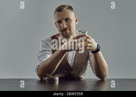 Stattlicher Mann mit Bart mit einem stilvollen Schnurrbart, in einem klassischen braunen Weste und weißem Hemd gekleidet sitzen am Tisch ist, Whiskey und Stockfoto