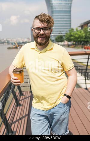 Bärtiger tausendjähriger Mann, der Kaffee zum Mitnehmen am frühen Morgen in einem sonnigen Park in der Nähe des Flusses in Belgrad hält Stockfoto