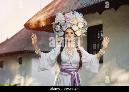 Schöne brünette Dame mit langen dunklen Haaren, in einem weißen ukrainischen authentischen Nationalkostüm und einem Kranz aus bunten Blumen gestikuliert während po Stockfoto